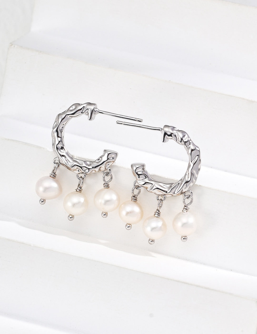 Pair of textured pearl hoop earrings in platinum silver, with three pearl drops on each hoop, displayed on a white block